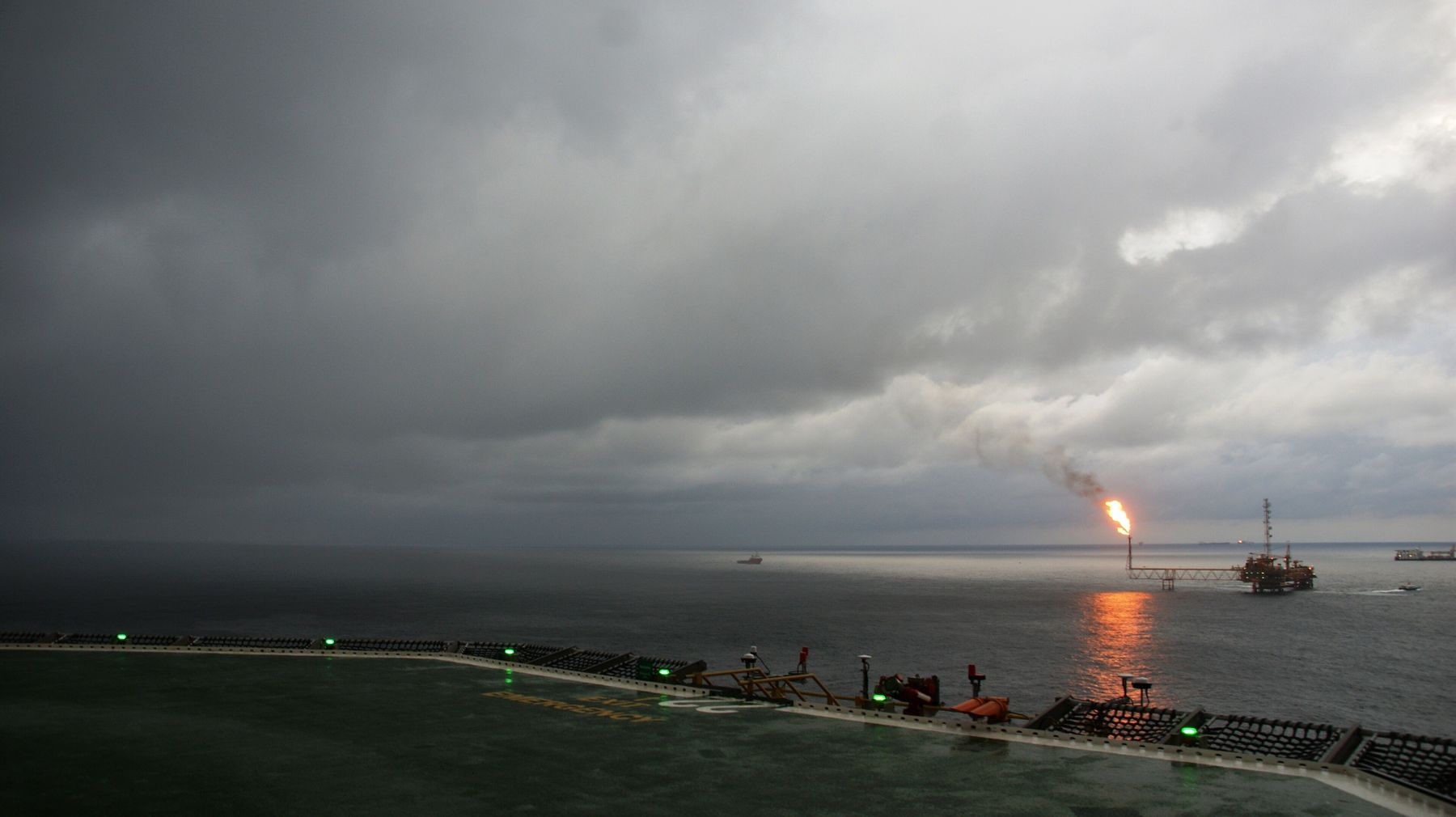 Heavy grey storm clouds over a dark, flat sea; on the horizon an offshore platform burns a bright gas flare.
