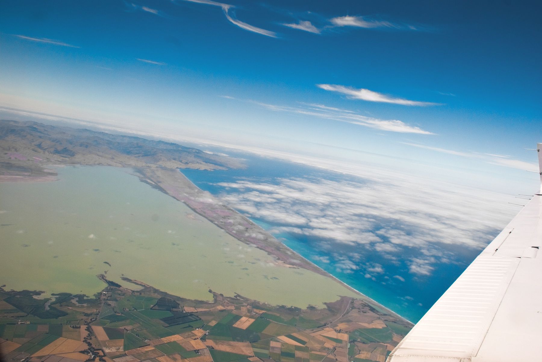 An aerial view past an aircraft wing: a broad greenish lagoon separated from the blue sea by a long thin strip of shore, farmland in the foreground.
