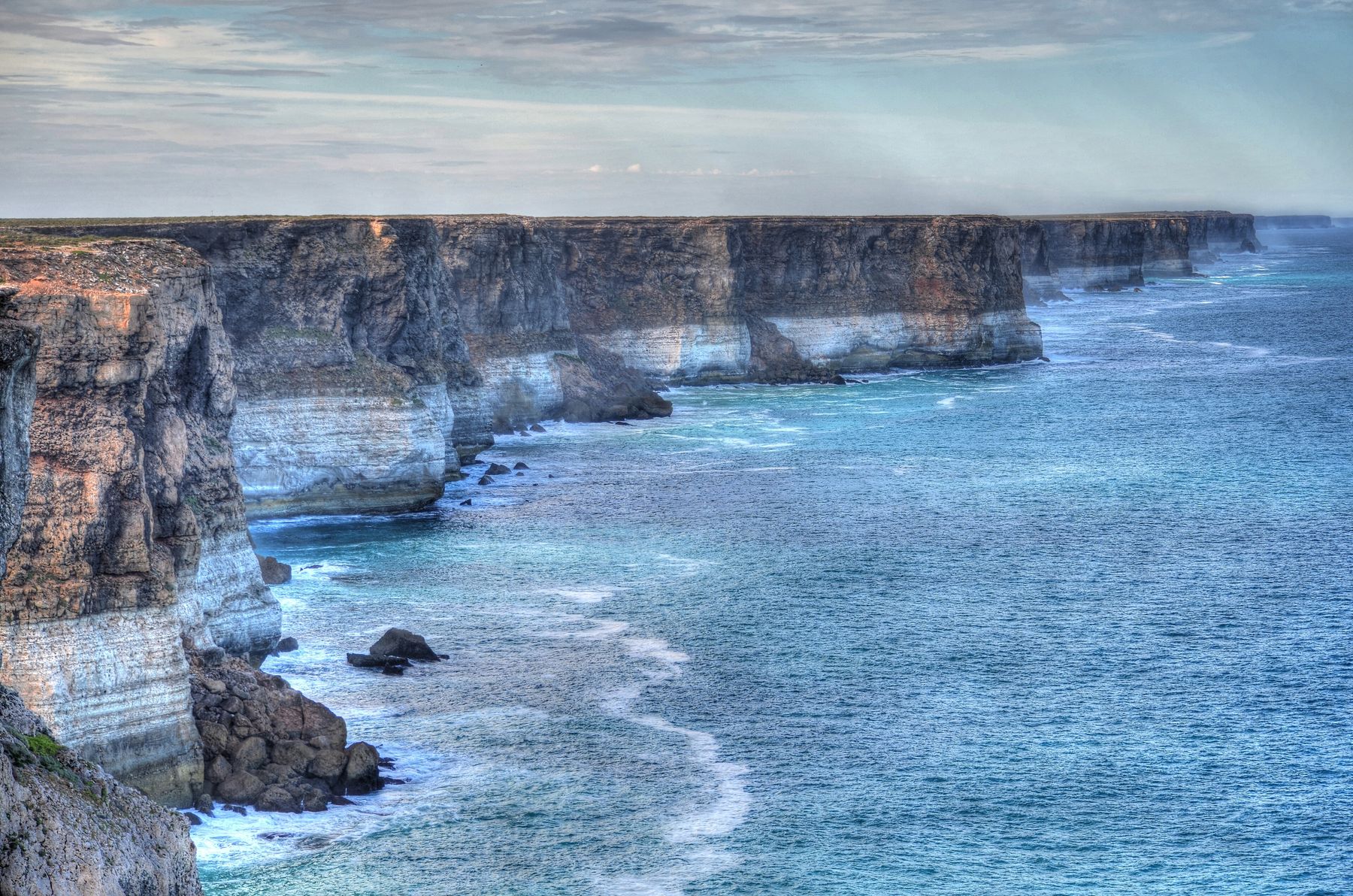 Banded cream and brown cliffs run in a wall along the coast while white surf breaks at their feet and on scattered rocks.