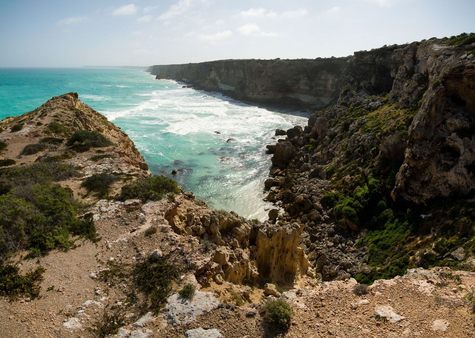 From a scrubby clifftop, a rocky cove opens to turquoise water and white surf, with a long line of cliffs stretching away to the left.