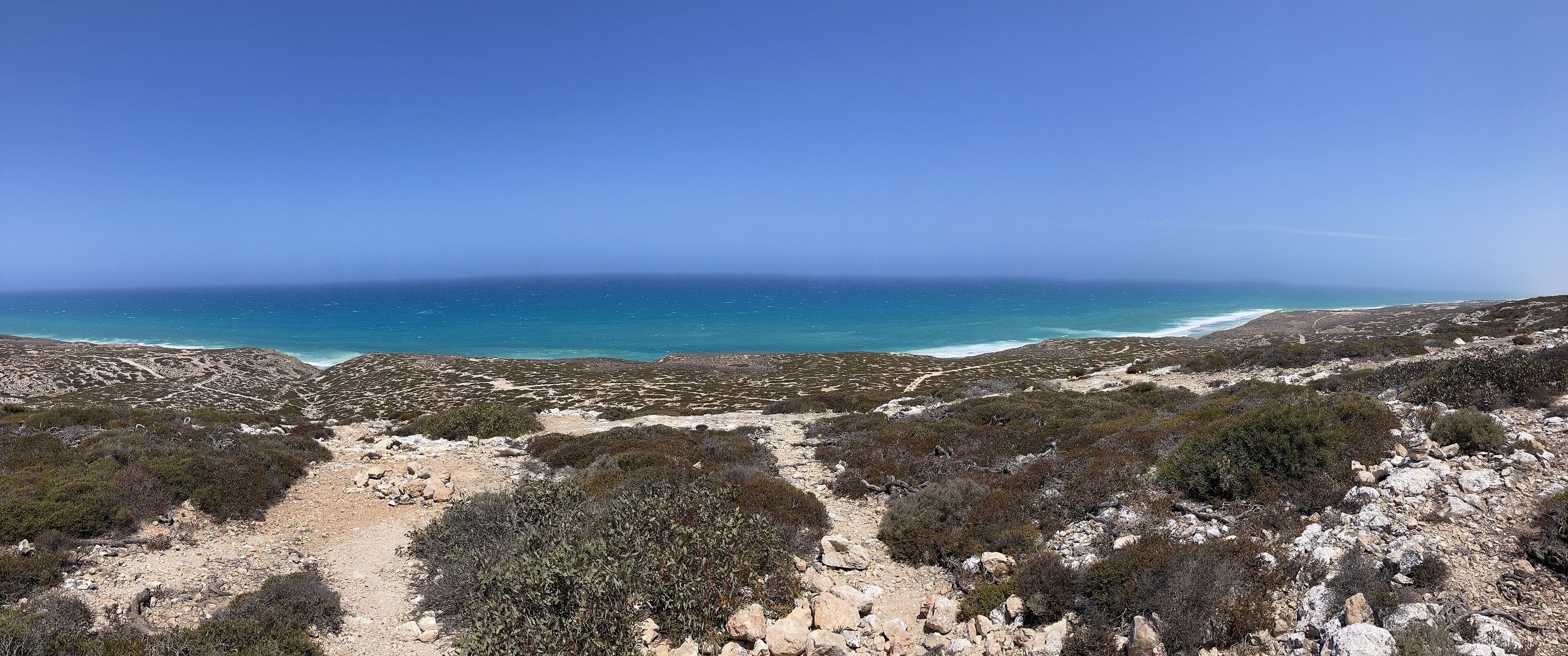 A wide view over low grey-green scrub and a sandy track to a long, flat blue sea line under an open sky.