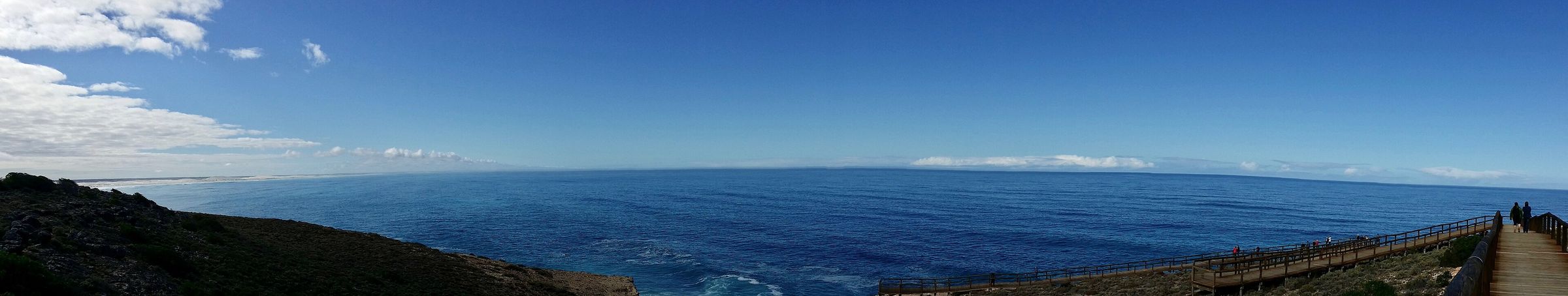 A very wide panorama: deep blue sea under a pale sky, with a low dark cliff line running along the right and a boardwalk at the lower right edge.