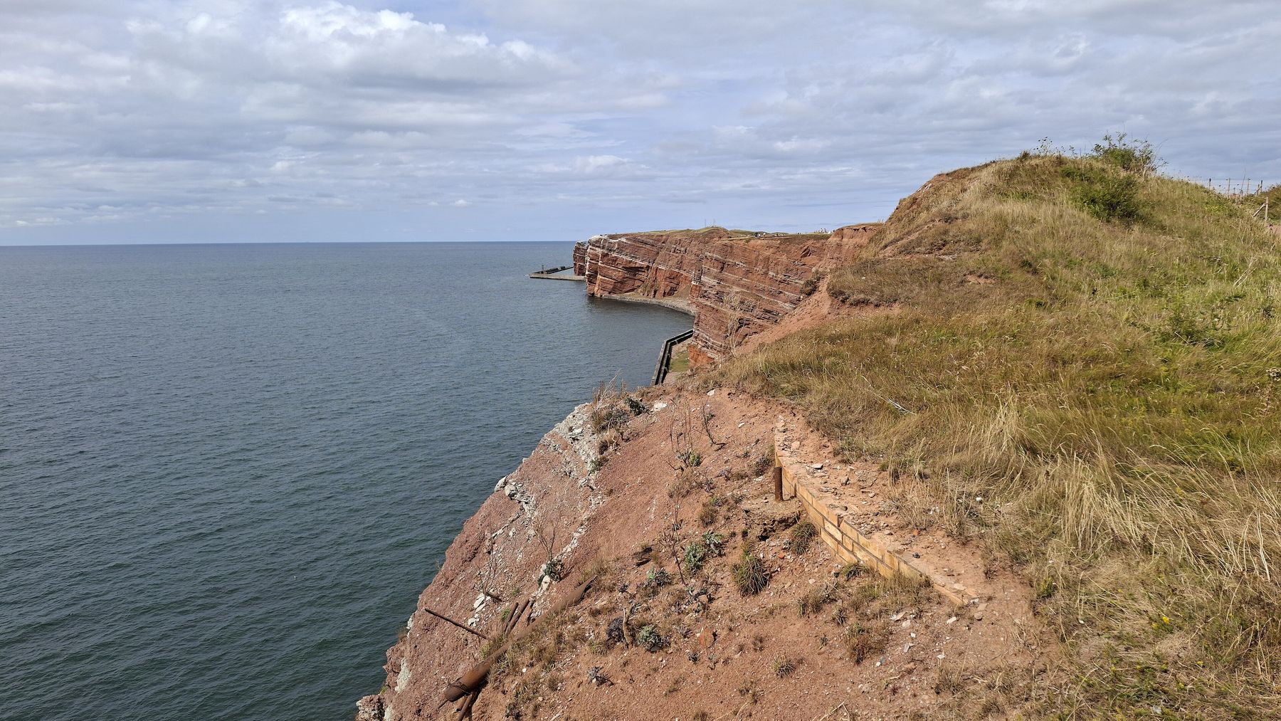 A grassy clifftop of red rock falls steeply to calm grey sea, the cliff edge curving away toward a distant headland.