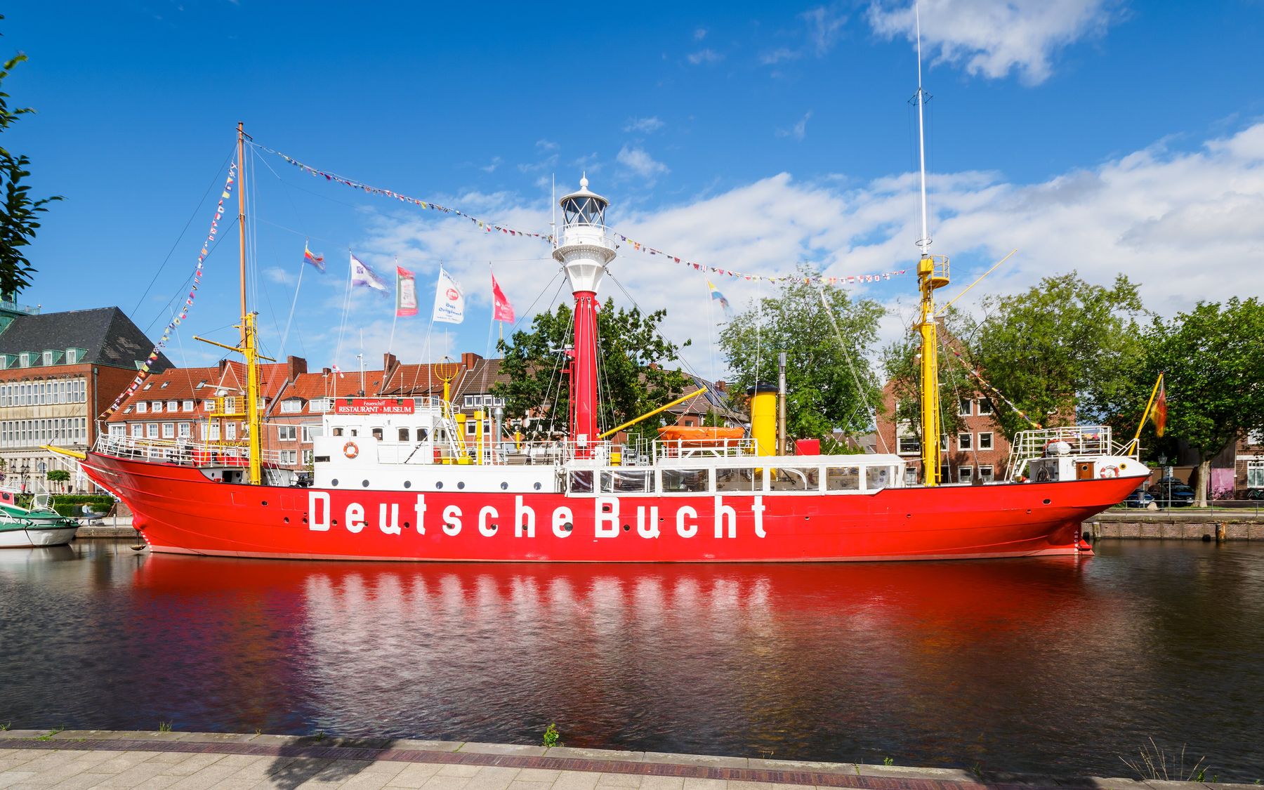 A red lightship with a white superstructure, a red lantern tower and the words 'Deutsche Bucht' on its hull, moored in still water in front of brick buildings.