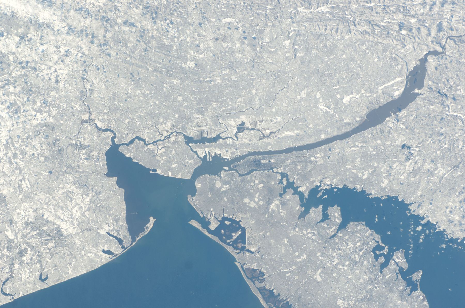 A snowy grey-white landscape seen from orbit with blue harbour water reaching inland; a long narrow beach runs along the lower edge where the open sea begins.