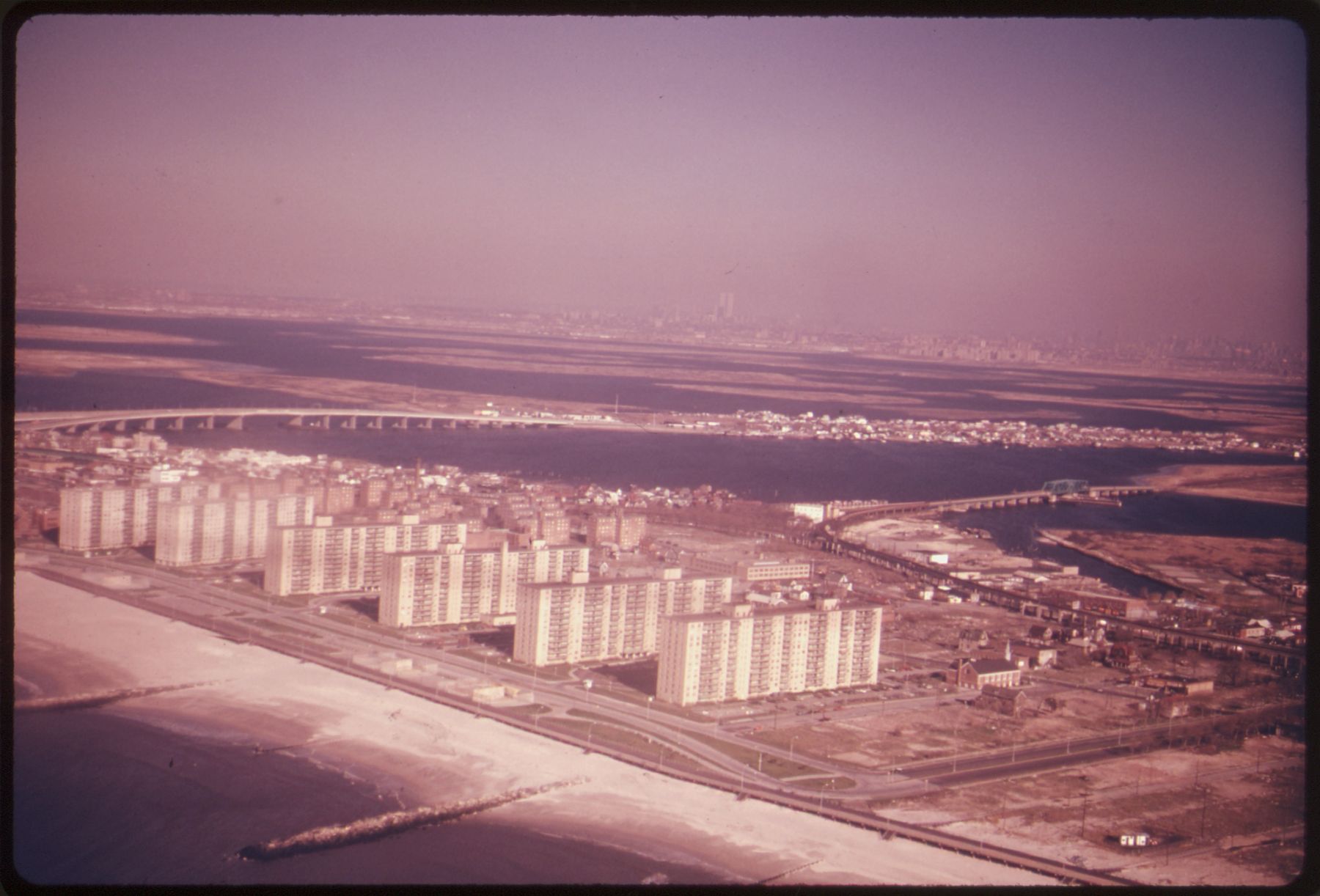 An aerial colour photograph with a faded pink cast: a sandy beach and rows of tall apartment blocks along the shore, with a bridge and water behind.