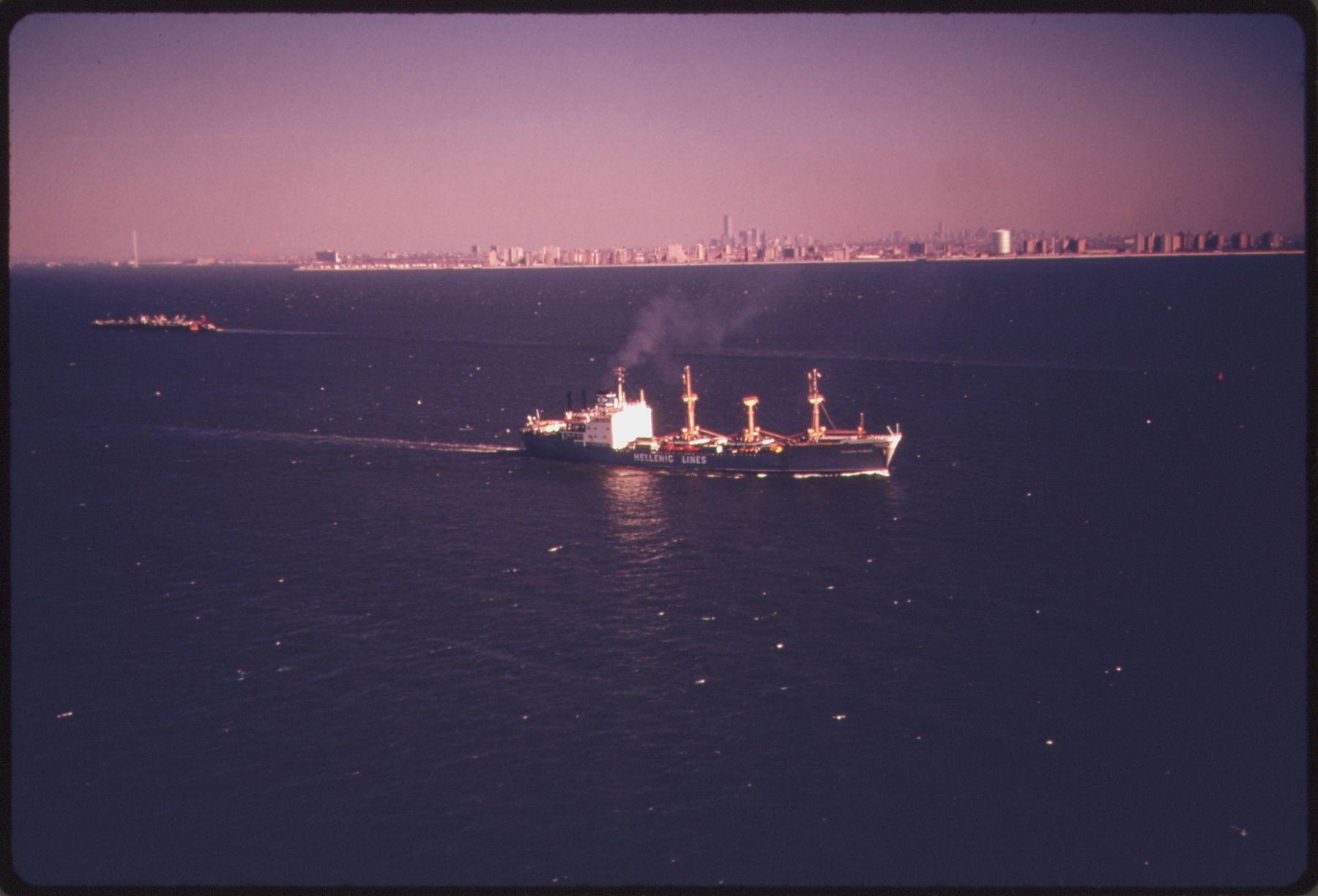 A cargo ship trailing smoke crosses dark open water, with a thin line of city towers on the far horizon.