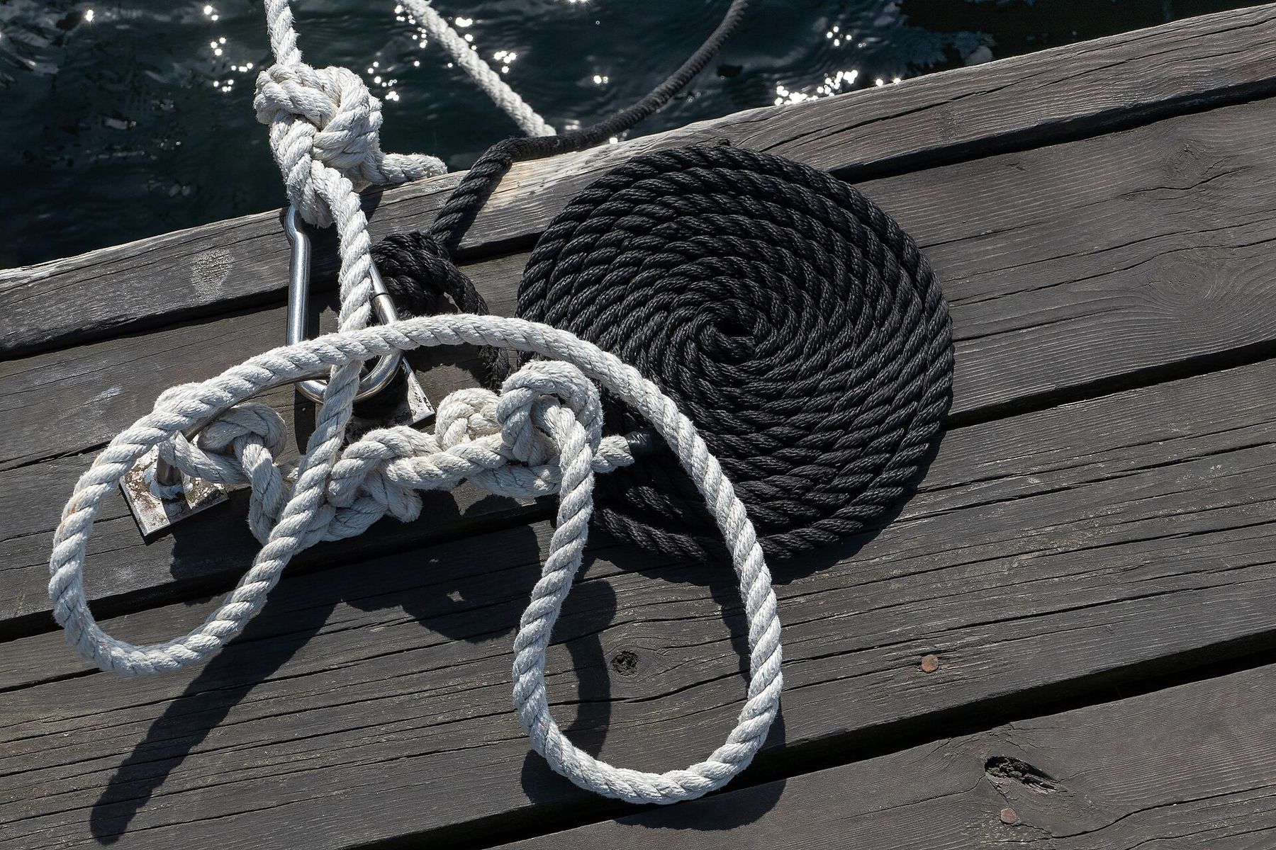 On grey jetty planks, a black rope lies in a tight flat spiral beside a white rope tied around a cleat, its slack falling in open loops.