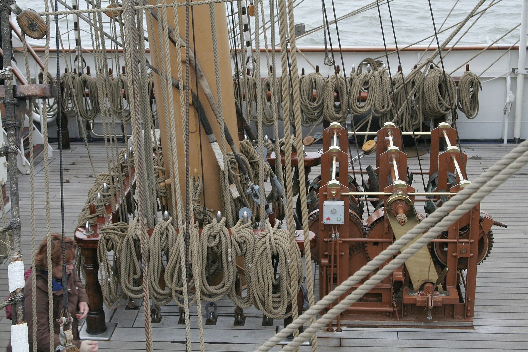 The deck of a tall ship crowded with vertical lines of rigging; dozens of rope coils hang in rows from pin rails around a red-painted winch.