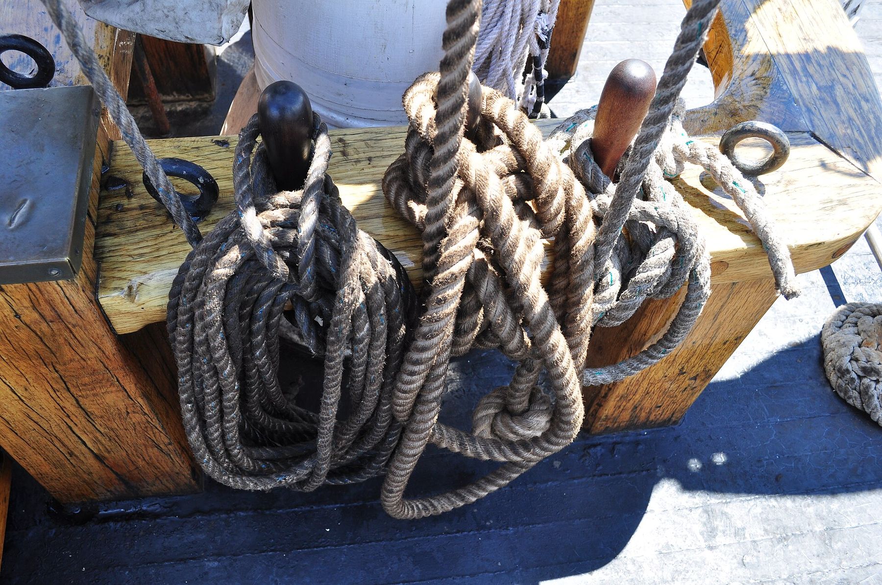 Coils of dark rope hanging from wooden belaying pins at the foot of a mast, with dark iron fittings and sunlit deck around them.